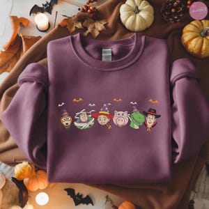 May include: A mauve sweatshirt featuring a Halloween-themed design with cartoon characters in costumes. The design includes characters like Woody, Buzz Lightyear, and others, each wearing a witch hat or wrapped in bandages. Small bat silhouettes are above the characters.