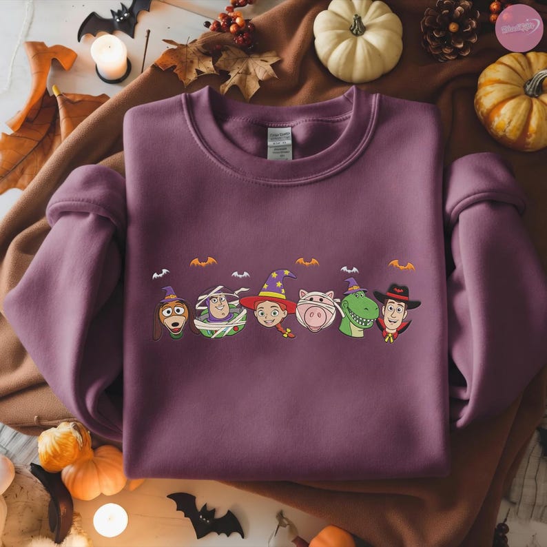 May include: A mauve sweatshirt featuring a Halloween-themed design with cartoon characters in costumes. The design includes characters like Woody, Buzz Lightyear, and others, each wearing a witch hat or wrapped in bandages. Small bat silhouettes are above the characters.