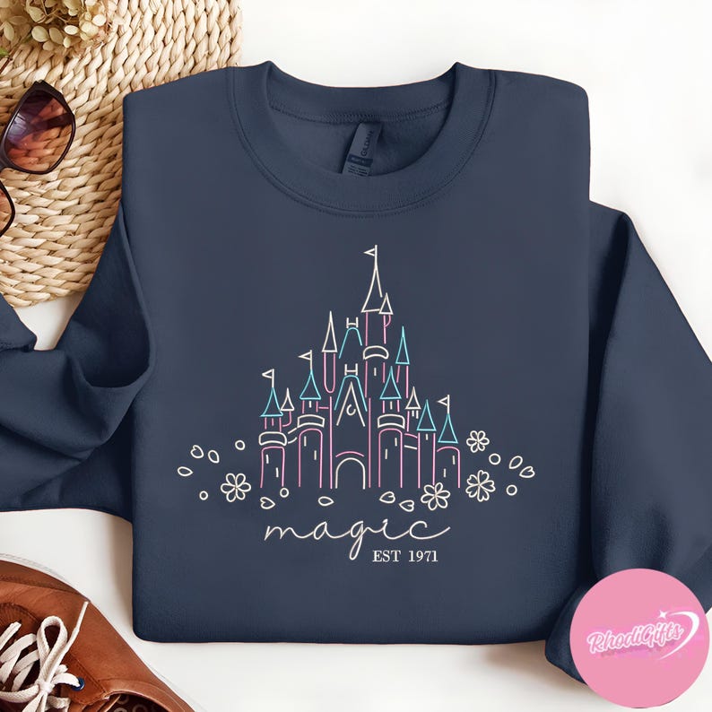 May include: Navy blue sweatshirt featuring a line drawing of a castle in pink and blue, with the word "magic" and "EST 1971" in white script. The design includes floral accents.
