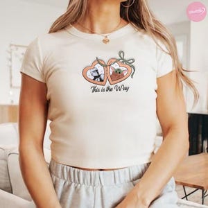 K&ouml;nnte beinhalten: Cremefarbenes T-Shirt mit herzf&ouml;rmigem Design. Das Design zeigt Illustrationen eines Charakters in wei&szlig;er R&uuml;stung und eines gr&uuml;nen Au&szlig;erirdischen mit dem Text "This is the Way". Das T-Shirt hat kurze &Auml;rmel und einen Rundhalsausschnitt.