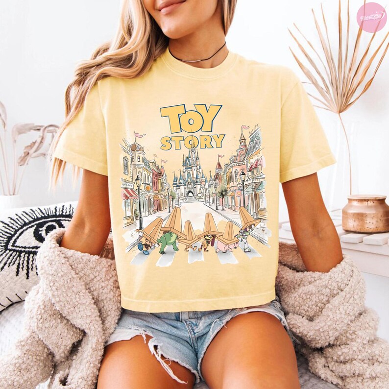 May include: Yellow short-sleeved t-shirt featuring the words "Toy Story" in a blue and yellow font. The graphic depicts a cartoon scene of characters crossing a street with buildings and a castle in the background.