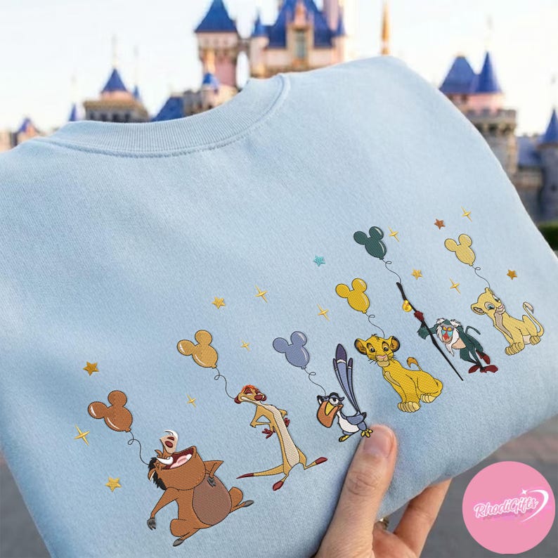 K&ouml;nnte beinhalten: Hellblaues Sweatshirt mit einer Stickerei von Figuren aus K&ouml;nig der L&ouml;wen. Simba, Timon und Pumbaa halten Mickey Mouse-Ballons, mit goldenen Sternen auf dem Stoff verstreut.