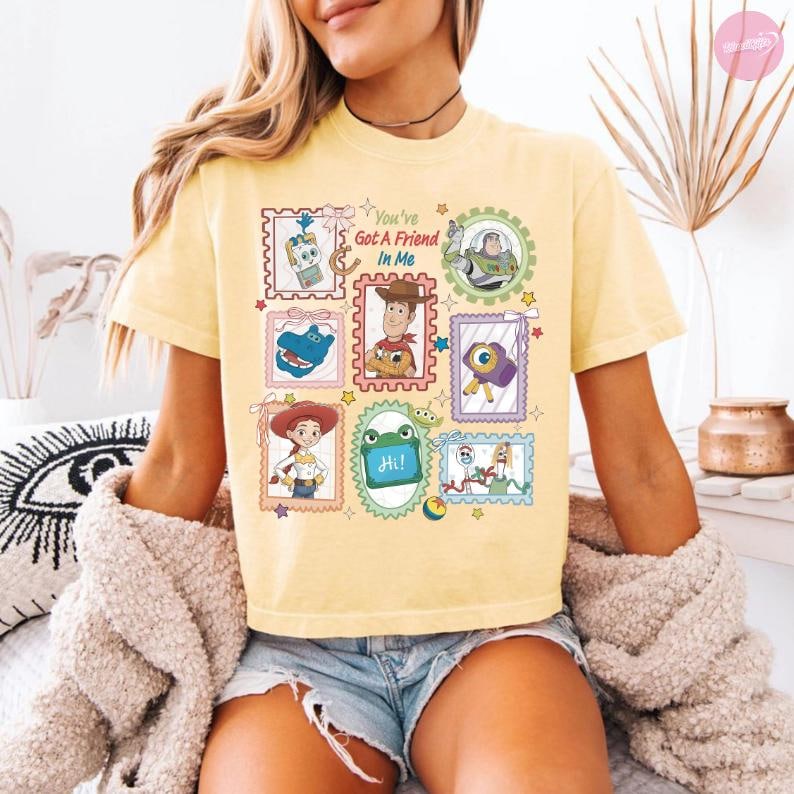 May include: A pale yellow t-shirt featuring a graphic print of various Toy Story characters arranged in postage stamp-style frames. The text "You've Got A Friend In Me" is printed in the center. Characters include Woody, Jessie, Buzz Lightyear, and Forky.
