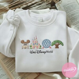 Peut inclure: Sweat-shirt blanc avec une broderie Walt Disney World. Le motif comprend les c&eacute;l&egrave;bres oreilles de Mickey, un ch&acirc;teau, une sph&egrave;re g&eacute;om&eacute;trique et un arbre. Les mots "Walt Disney World" sont brod&eacute;s en dessous.