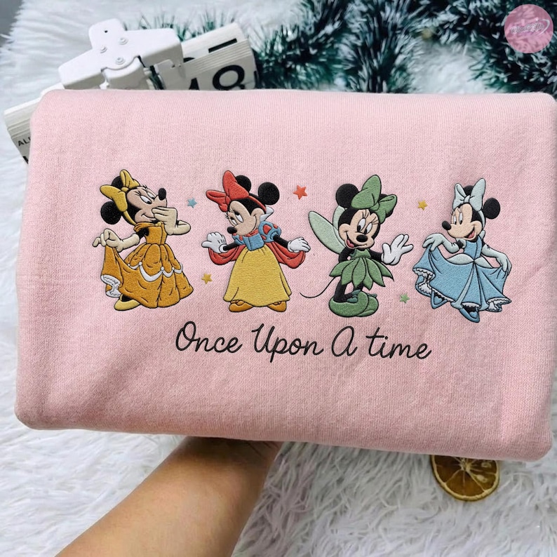 K&ouml;nnte beinhalten: Hellrosa Sweatshirt mit vier Minnie Mouse-Figuren in Prinzessinnen-Outfits. Der Schriftzug "Once Upon A Time" ist darunter gestickt. Das Sweatshirt hat eine hellrosa Farbe.