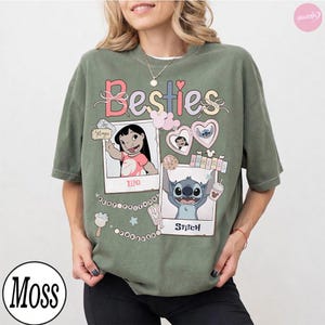 Puede incluir: Camiseta verde oliva con la palabra "Besties" en colores pastel. La camiseta presenta im&aacute;genes estilo polaroid de Lilo y Stitch, junto con gr&aacute;ficos de corazones, helados y palomitas de ma&iacute;z. La camiseta es de corte holgado.