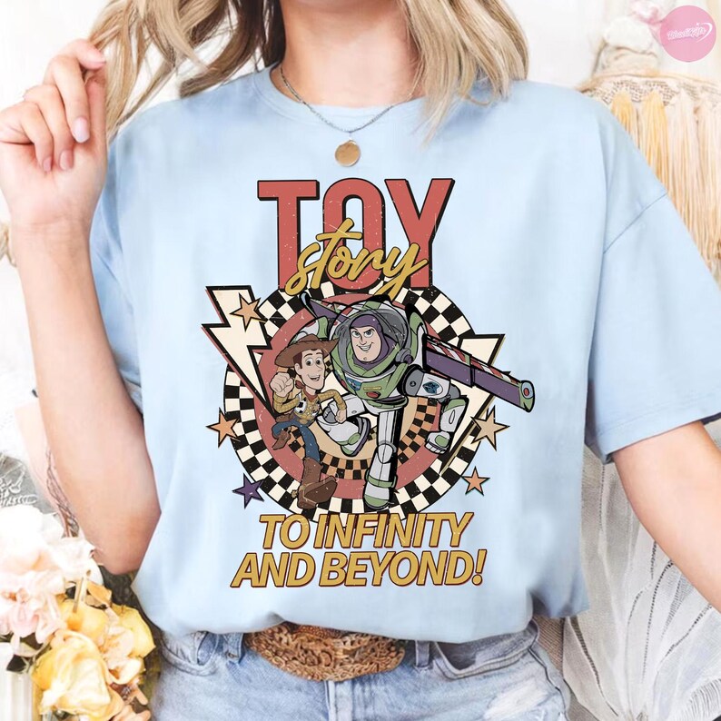May include: Pale blue t-shirt with a graphic of Woody and Buzz Lightyear from Toy Story. The design includes the words "Toy Story" and "To Infinity and Beyond!" in a retro style, with a checkered background and star accents.