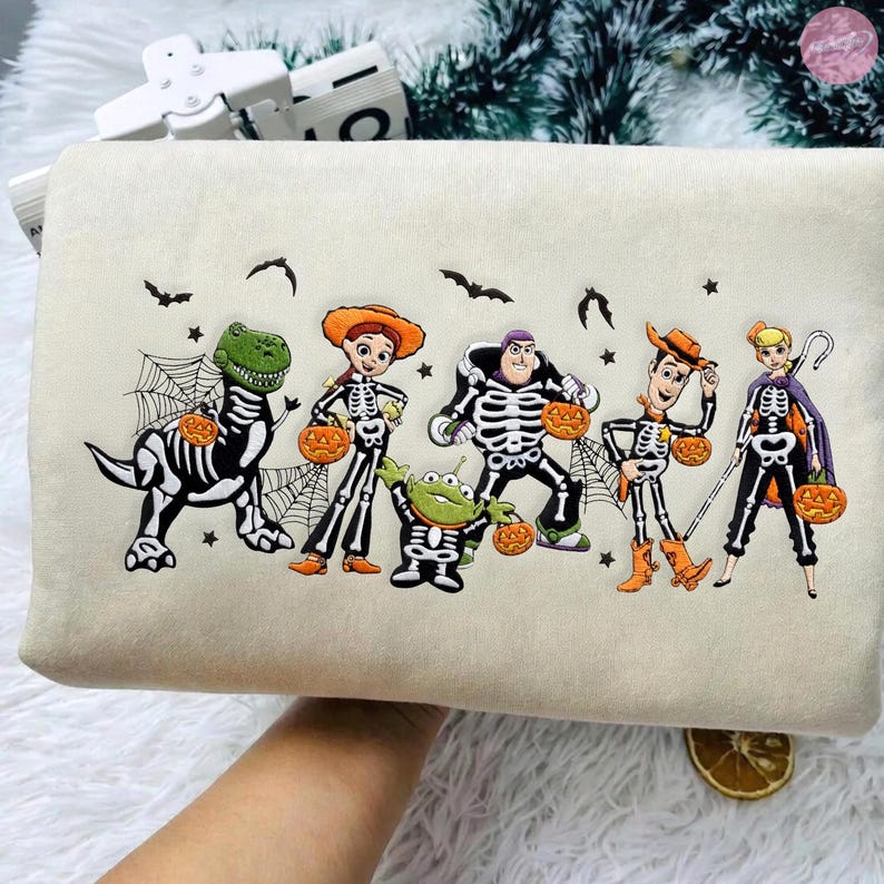 May include: A cream-colored sweatshirt featuring an embroidered graphic of five cartoon characters dressed in skeleton costumes for Halloween. Each character holds a small orange pumpkin, surrounded by black bats, stars, and spider webs.