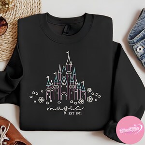 May include: Black crewneck sweatshirt featuring a castle design in white, pink, and teal. The word "magic" is written in cursive below the castle, with "EST 1971" underneath. The design includes floral accents.