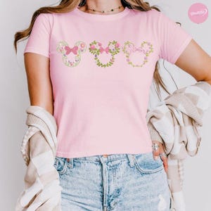 May include: A light pink t-shirt featuring three floral Mickey Mouse head designs. Each design is a different floral arrangement with a bow. The shirt is made of a soft material and is suitable for casual wear.