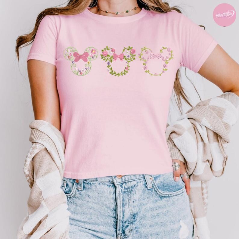 May include: A light pink t-shirt featuring three floral Mickey Mouse head designs. Each design is a different floral arrangement with a bow. The shirt is made of a soft material and is suitable for casual wear.