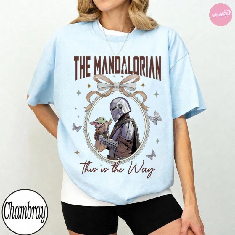 May include: Light blue t-shirt featuring a graphic from The Mandalorian. The design includes the text "THE MANDALORIAN" above an illustration of the Mandalorian holding Grogu, with the phrase "This is the Way" below. Butterflies and stars surround the central image.