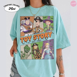 May include: A mint green t-shirt featuring a graphic print of Toy Story characters dressed in Halloween costumes. The design includes Buzz Lightyear, Woody, Jessie, Rex, Slinky Dog, and Bo Peep, with the text Toy Story Est. 1995 in orange.