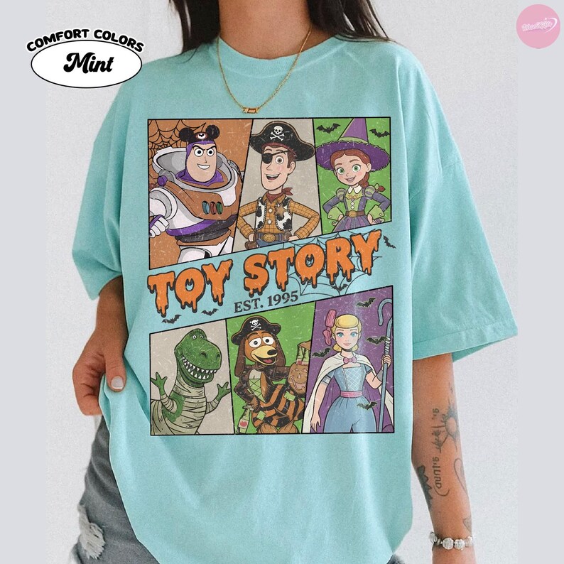 May include: A mint green t-shirt featuring a graphic print of Toy Story characters dressed in Halloween costumes. The design includes Buzz Lightyear, Woody, Jessie, Rex, Slinky Dog, and Bo Peep, with the text Toy Story Est. 1995 in orange.