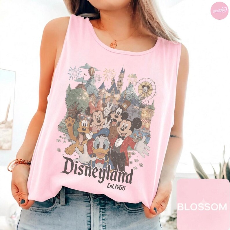 May include: A light pink sleeveless tank top featuring a graphic print of cartoon characters including Mickey Mouse, Minnie Mouse, Donald Duck, and Pluto in front of a castle. The text reads Disneyland Est. 1955 in a dark brown vintage-style font.