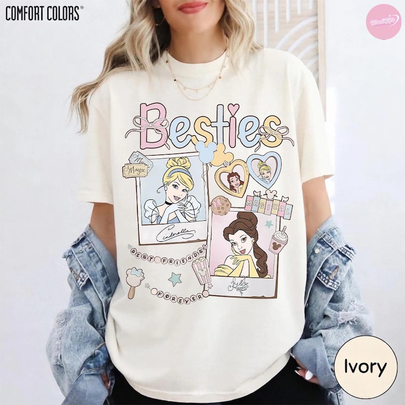 Puede incluir: Camiseta color marfil con la palabra "Besties" en colores pastel. La camiseta presenta ilustraciones de princesas de Disney en marcos estilo polaroid, con las palabras "Cinderella" y "Belle" debajo. Hecha de material Comfort Colors.