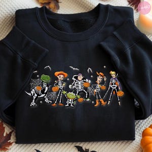 May include: A black crewneck sweatshirt featuring a graphic print of five animated characters dressed in skeleton costumes for Halloween. Each character holds a small orange jack-o'-lantern, surrounded by white spiderwebs, stars, and bats.