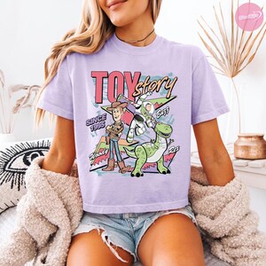 Op de afbeelding: Lichtpaars T-shirt met een afbeelding van Woody, Buzz Lightyear en Rex uit Toy Story. Het ontwerp bevat de woorden "Toy Story" en "Since 1995" in een retrostijl.