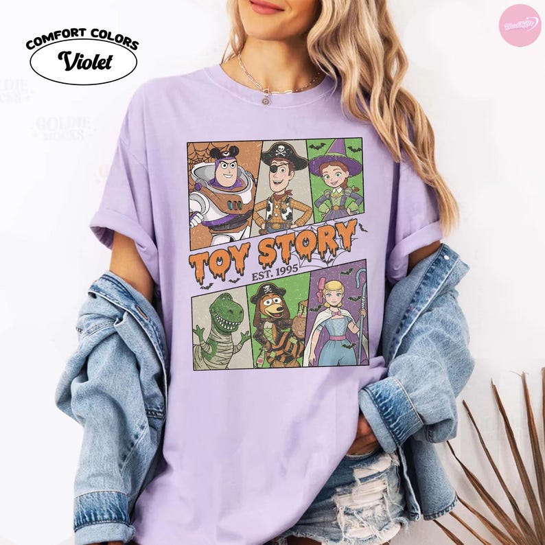 May include: A violet short-sleeve t-shirt featuring a graphic print of Toy Story characters in Halloween costumes. The design includes Buzz Lightyear, Woody, Jessie, Rex, Slinky Dog, and Bo Peep with the text Toy Story Est. 1995.