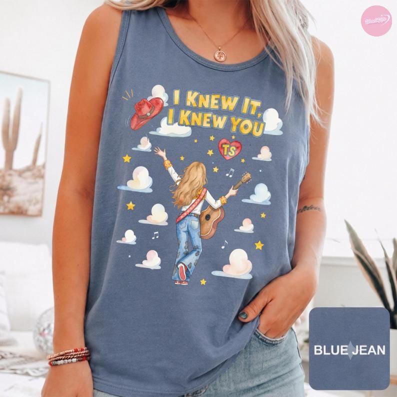 May include: A blue jean colored tank top featuring a graphic illustration of a person with long blonde hair playing an acoustic guitar. The text reads I KNEW IT, I KNEW YOU with a red cowboy hat, a heart with TS, and yellow stars and white clouds.