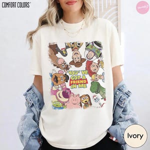 May include: Ivory-colored Comfort Colors t-shirt featuring a colorful graphic of characters from the movie Toy Story. The graphic includes the text "YOU'VE GOT A FRIEND IN ME". The shirt is a crew neck and has short sleeves.