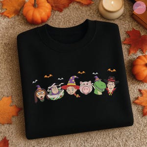 May include: Black sweatshirt featuring a Halloween-themed design with cartoon character faces, including a dog, Buzz Lightyear, Jessie, a pig, a dinosaur, and Woody. Bats are above the characters. The sweatshirt is surrounded by pumpkins and fall leaves.