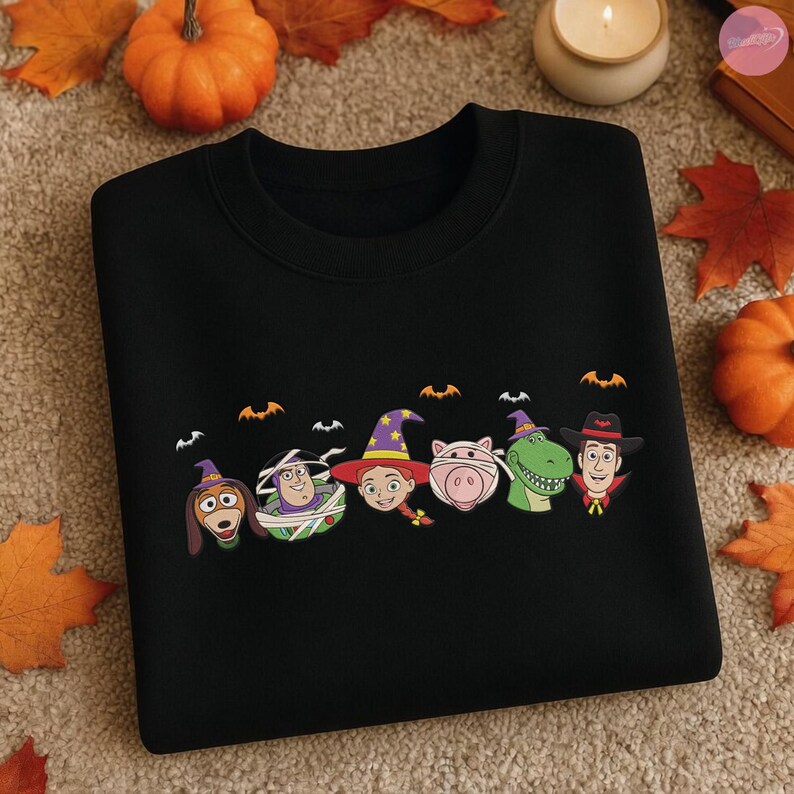 May include: Black sweatshirt featuring a Halloween-themed design with cartoon character faces, including a dog, Buzz Lightyear, Jessie, a pig, a dinosaur, and Woody. Bats are above the characters. The sweatshirt is surrounded by pumpkins and fall leaves.