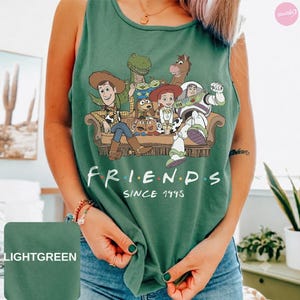 May include: A sage green sleeveless tank top featuring a graphic print of various Toy Story characters sitting on a couch. Below the illustration, the text reads "FRIENDS SINCE 1995" in white capital letters. The shirt has a round neckline.