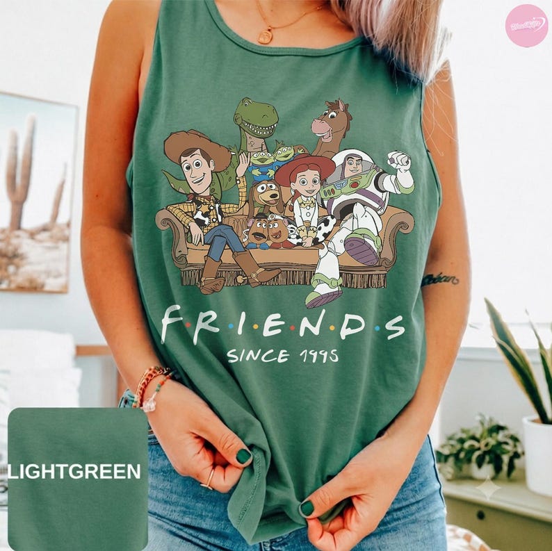 May include: A sage green sleeveless tank top featuring a graphic print of various Toy Story characters sitting on a couch. Below the illustration, the text reads "FRIENDS SINCE 1995" in white capital letters. The shirt has a round neckline.