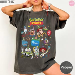May include: A charcoal gray short-sleeve t-shirt featuring a colorful graphic print with the text "BIRTHDAY STORY" and "Name". The design includes various cartoon characters wearing party hats, a red balloon with the number 5, and confetti.