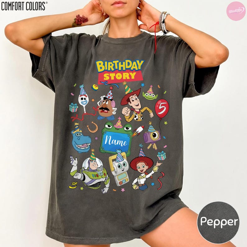 May include: A charcoal gray short-sleeve t-shirt featuring a colorful graphic print with the text "BIRTHDAY STORY" and "Name". The design includes various cartoon characters wearing party hats, a red balloon with the number 5, and confetti.