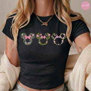 May include: Black t-shirt featuring three floral Mickey Mouse head designs. Each design includes a pink bow and is surrounded by flowers and greenery. The shirt has short sleeves and a crew neck.