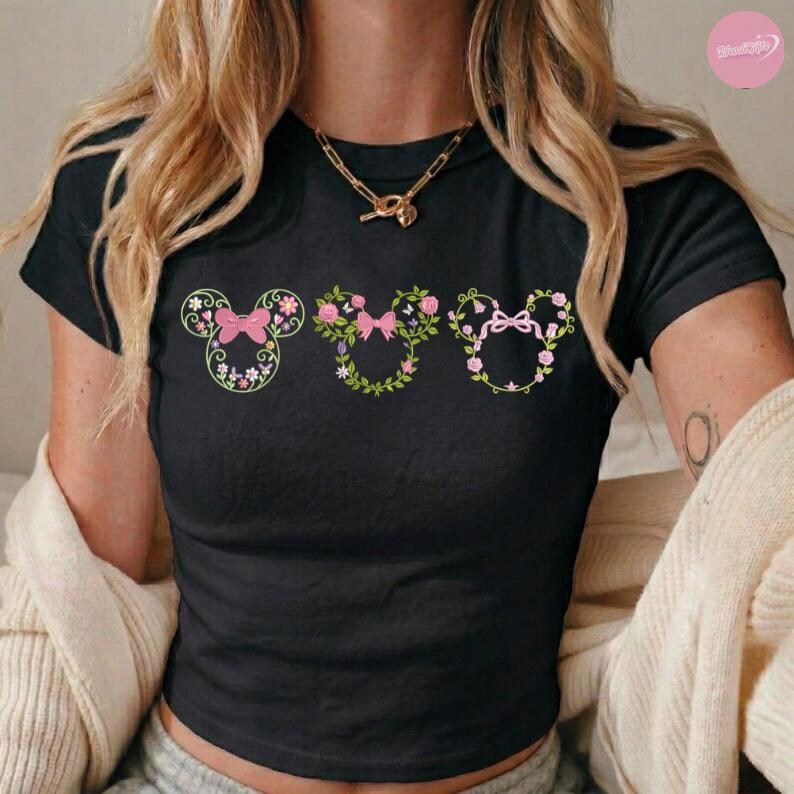May include: Black t-shirt featuring three floral Mickey Mouse head designs. Each design includes a pink bow and is surrounded by flowers and greenery. The shirt has short sleeves and a crew neck.