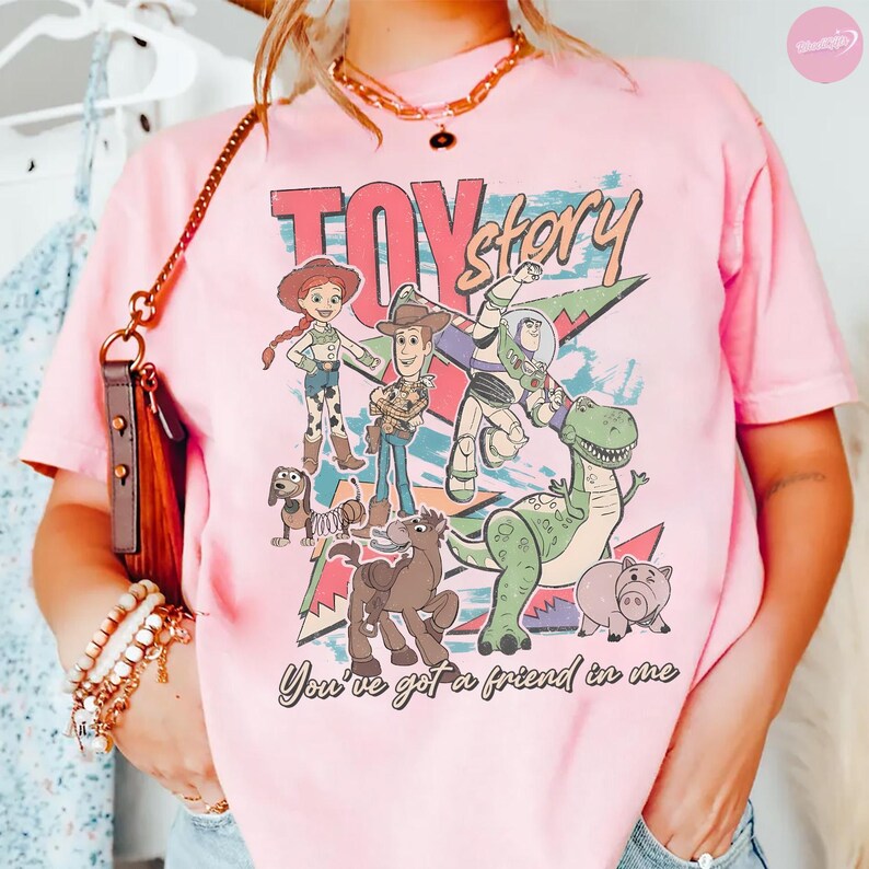 Puede incluir: Camiseta rosa claro con el texto "Toy Story" en una fuente retro, con personajes de dibujos animados como Woody y Buzz Lightyear. La frase "You've got a friend in me" est&aacute; impresa debajo.