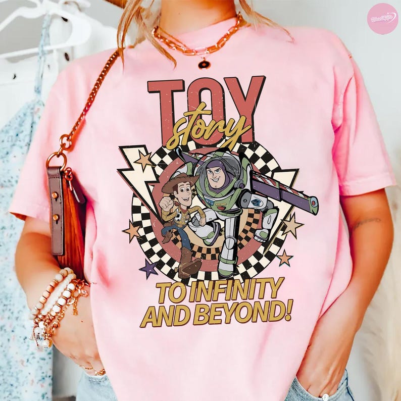 May include: Pale pink t-shirt with a graphic of Woody and Buzz Lightyear from Toy Story. The design includes the words "TOY STORY" and "TO INFINITY AND BEYOND!" in a retro style. The shirt has a crew neck and short sleeves.