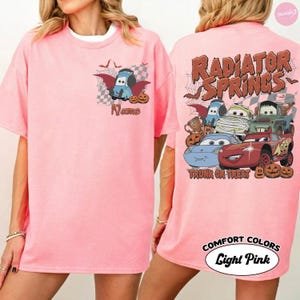 May include: A light pink t-shirt featuring a small graphic on the front with a blue car character, pumpkins, and the word Name. The back displays a large graphic with multiple car characters, pumpkins, spiderwebs, and the text Radiator Springs Trunk or Treat.