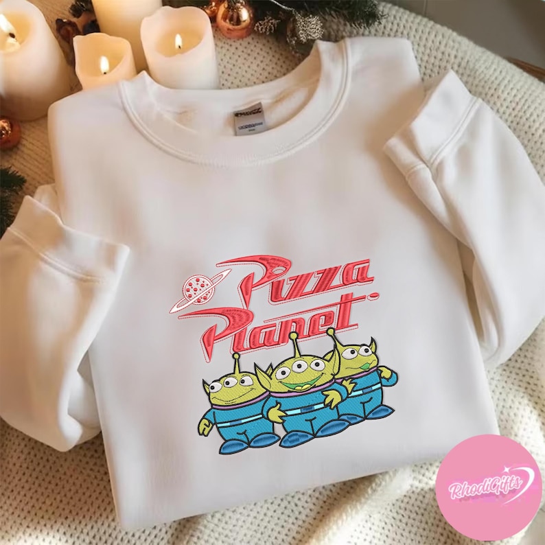 May include: White sweatshirt featuring the Pizza Planet logo in red with a planet graphic. Below the logo are three green aliens in blue uniforms. The sweatshirt is a crew neck style.