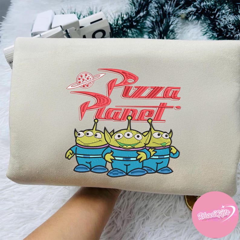 May include: A beige clutch bag with a red embroidered 'Pizza Planet' logo and a planet graphic. Below the logo are three green aliens in blue jumpsuits. The bag is rectangular in shape.