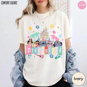 Pu&ograve; includere: T-shirt color avorio con la scritta "Disneyland" in colori pastello, con personaggi dei cartoni animati e una grafica del castello. La maglietta ha girocollo e maniche corte. Il marchio Comfort Colors &egrave; visibile in alto a sinistra.