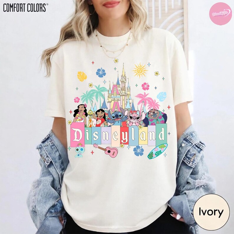Pu&ograve; includere: T-shirt color avorio con la scritta "Disneyland" in colori pastello, con personaggi dei cartoni animati e una grafica del castello. La maglietta ha girocollo e maniche corte. Il marchio Comfort Colors &egrave; visibile in alto a sinistra.