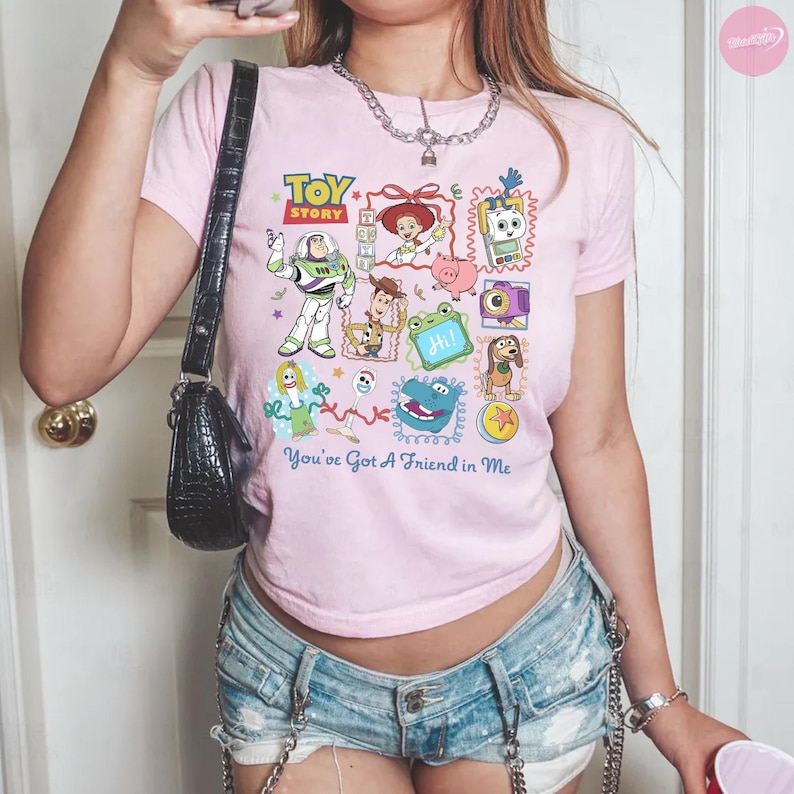 May include: A light pink t-shirt featuring a colorful graphic print of Toy Story characters including Buzz Lightyear, Woody, Jessie, and others, with the text "TOY STORY" and "You've Got A Friend in Me" printed in blue and red on the front.