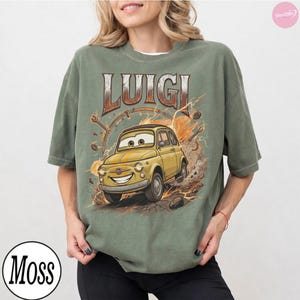 May include: Olive green t-shirt featuring a cartoon image of Luigi, a yellow car from the movie Cars. The word "Luigi" is printed above the car in a brown and orange font. The shirt has a crew neck and short sleeves.