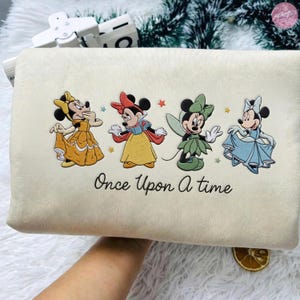 K&ouml;nnte beinhalten: Cremefarbene rechteckige Tasche mit gestickten Bildern von vier Zeichentrickfiguren in farbenfrohen Kleidern. Der Schriftzug "Once Upon a Time" steht in schwarzer Schrift darunter. Die Tasche wird von einer Person gehalten.