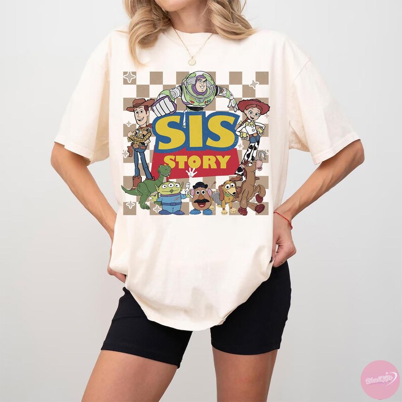 May include: Cream-colored t-shirt featuring a graphic of characters from the animated film Toy Story. The design includes Buzz Lightyear, Woody, Jessie, and other characters, with the text "SIS STORY" in yellow and red. The background has a checkered pattern.