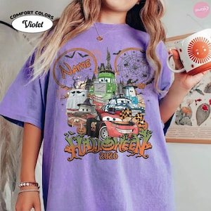 May include: A violet t-shirt featuring a graphic of animated cars in Halloween costumes, including a red race car with the number 95, a green truck, and a ghost car. The design includes spider webs, a castle, pumpkins, and the text Name Halloween 2026.