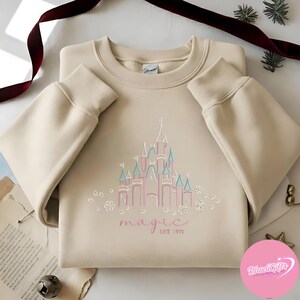 May include: A beige sweatshirt featuring a pink and blue castle design with the word "magic" and "EST 1971" in a script font. The design is surrounded by small white flowers. The sweatshirt has long sleeves with ribbed cuffs.