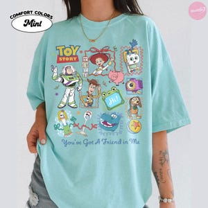 May include: A mint green short-sleeve t-shirt featuring a colorful graphic print of Toy Story characters including Buzz Lightyear, Woody, Jessie, and Forky. The design includes the text "TOY STORY" and "You've Got A Friend in Me" in blue lettering.