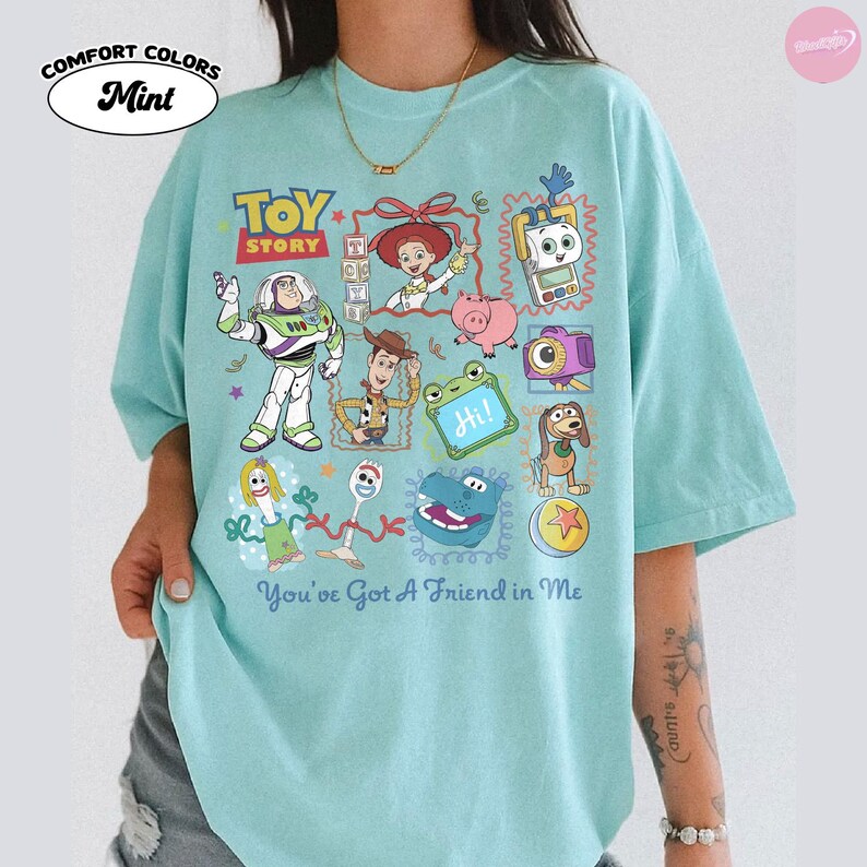 May include: A mint green short-sleeve t-shirt featuring a colorful graphic print of Toy Story characters including Buzz Lightyear, Woody, Jessie, and Forky. The design includes the text "TOY STORY" and "You've Got A Friend in Me" in blue lettering.