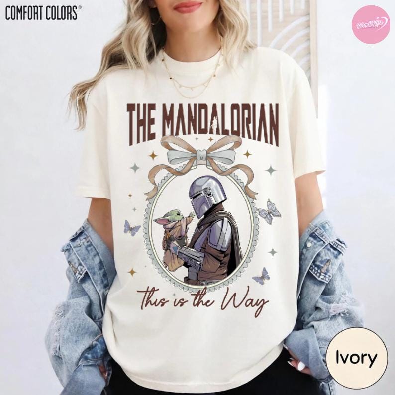 May include: Ivory t-shirt with a graphic of The Mandalorian and Grogu. The text "The Mandalorian" is above the image, and "This is the Way" is below. The design includes a bow, stars, and butterflies. The t-shirt is ivory in colour.
