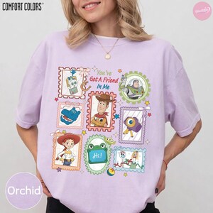 May include: A light purple t-shirt featuring a graphic print of various characters from the Toy Story franchise arranged in postage stamp-style frames. The text "You've Got A Friend In Me" is printed in the center, surrounded by colorful illustrations.
